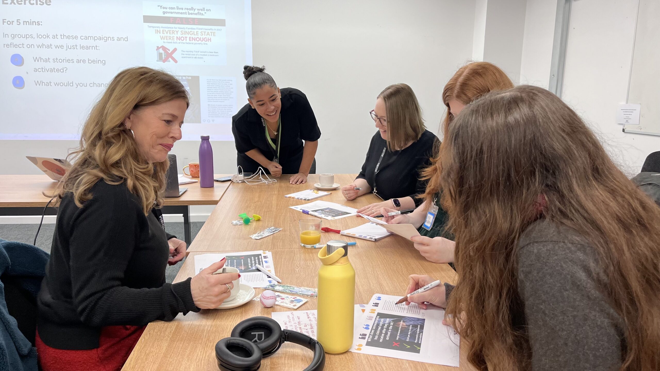 in person training session with Heard, facilitated by team member Makaela. 4 people sit around the table with her, with pens, headphones, papers. There is a screen projection behind them